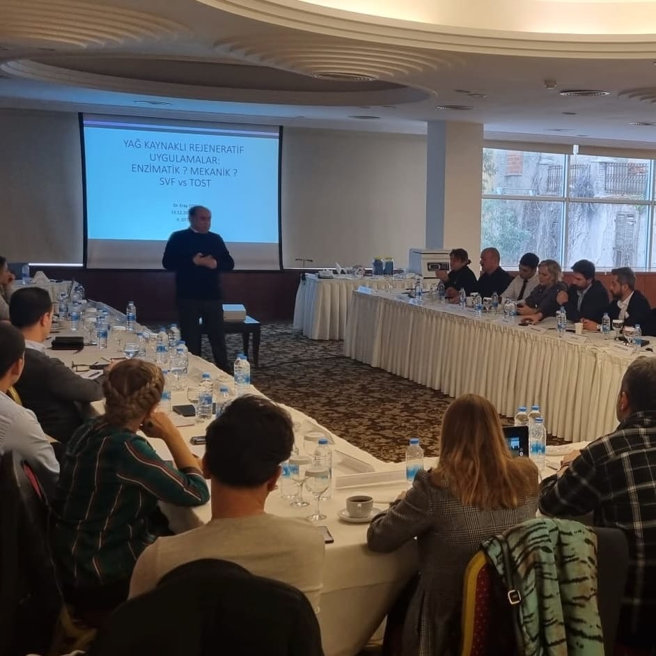 Copcu lecturing to a seated course beside a slide comparing enzymatic and mechanical SVF with TOST