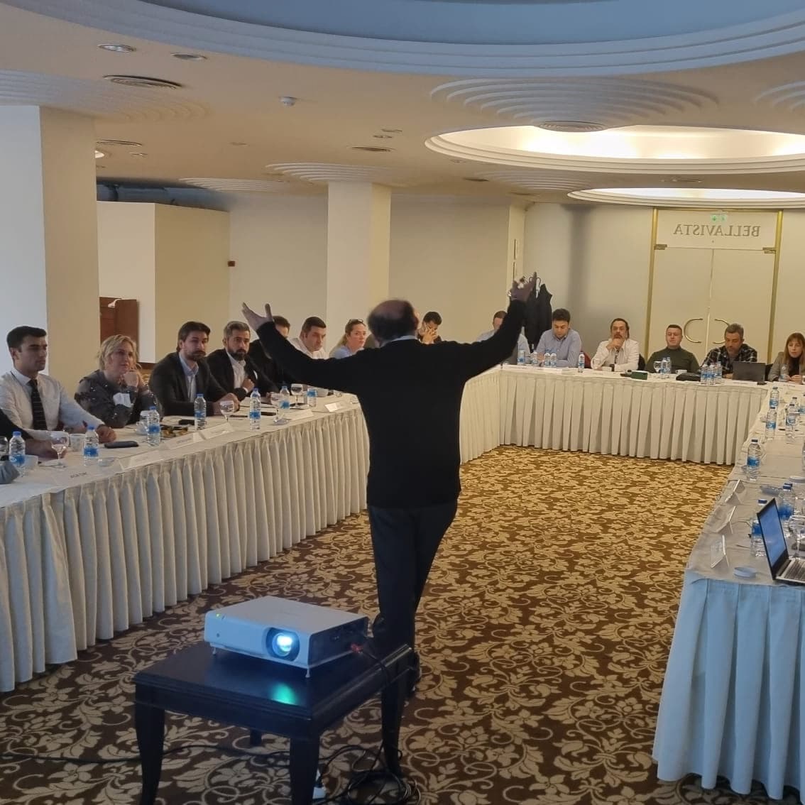 Copcu addressing delegates seated around a U-shaped table, arms open mid-sentence